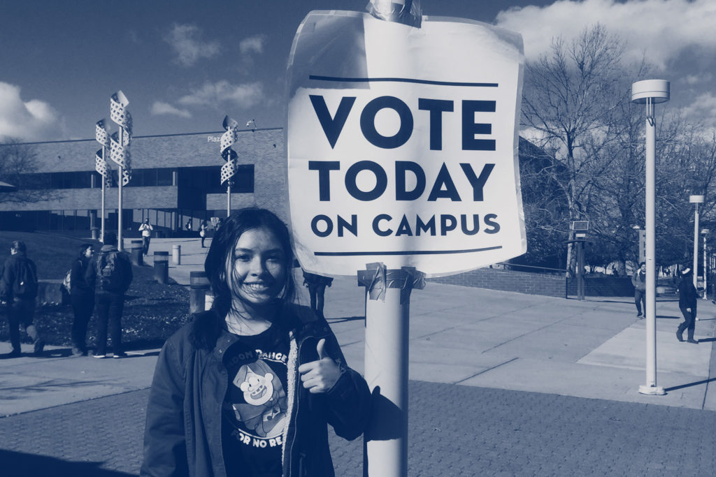 New Era organizer smiles and gives a thumbs up next to a sign that says Vote Today on Campus